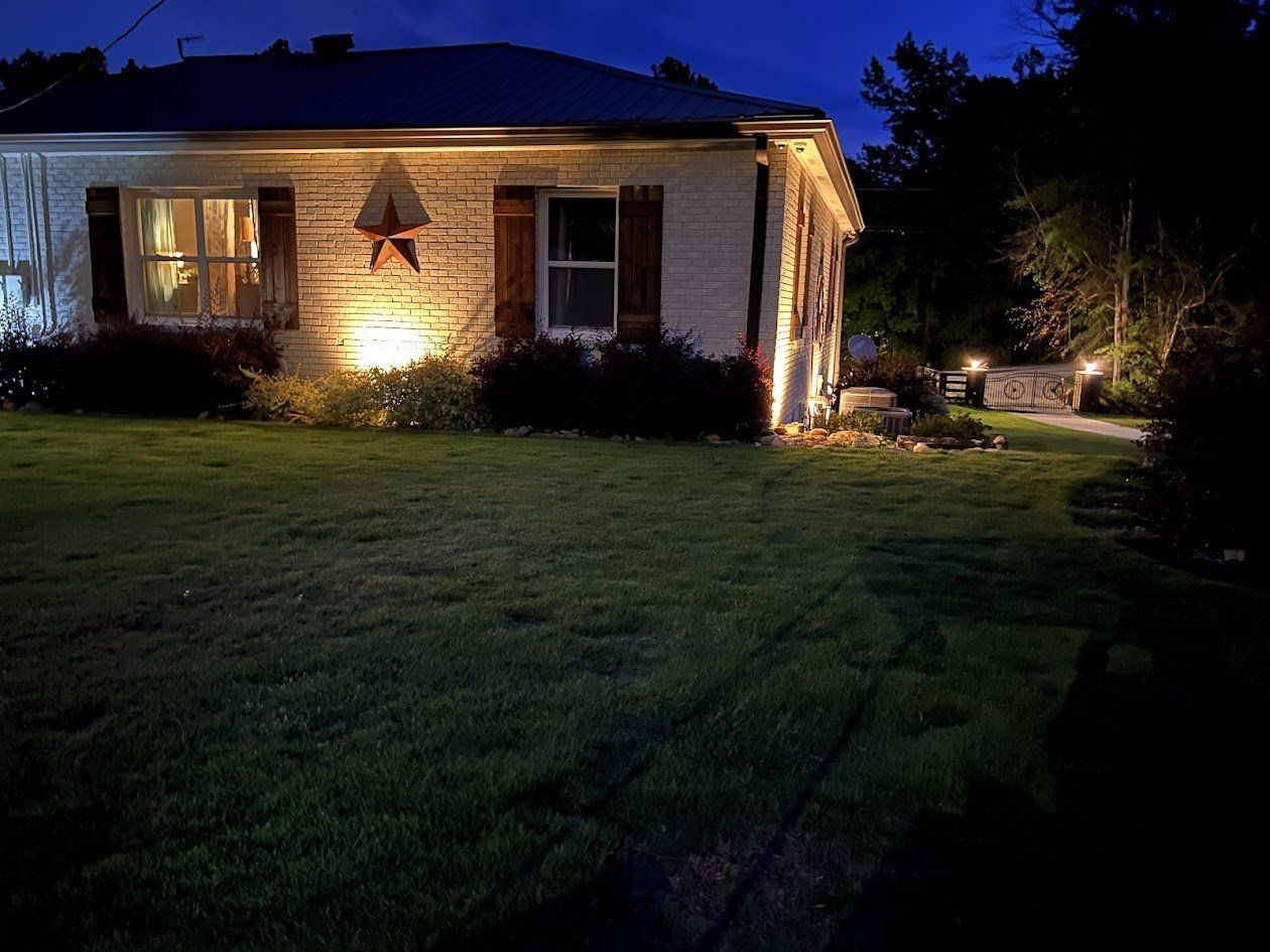 Side of a brick home uplit at night with lighted gate pillars in the background